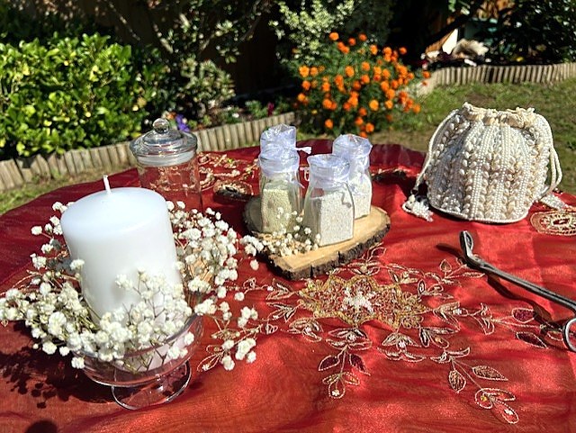 Four small jars of pale sand tied with sheer white fabric, a larger glass jar and a white pillar candle ringed with baby's breath, arranged on an embroidered crimson cloth outdoors