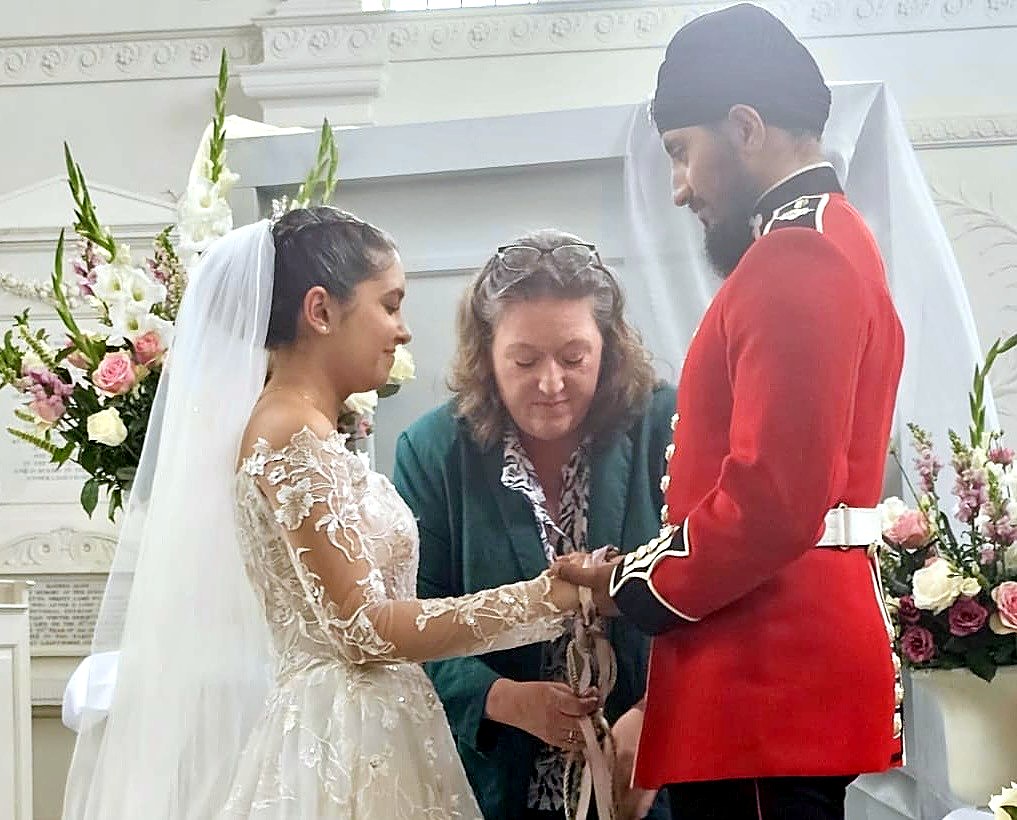 A celebrant binding the clasped hands of a bride and a groom in a scarlet ceremonial tunic with pale ribbon